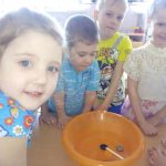 Smiling children stand next to a bowl of water with small objects floating in it